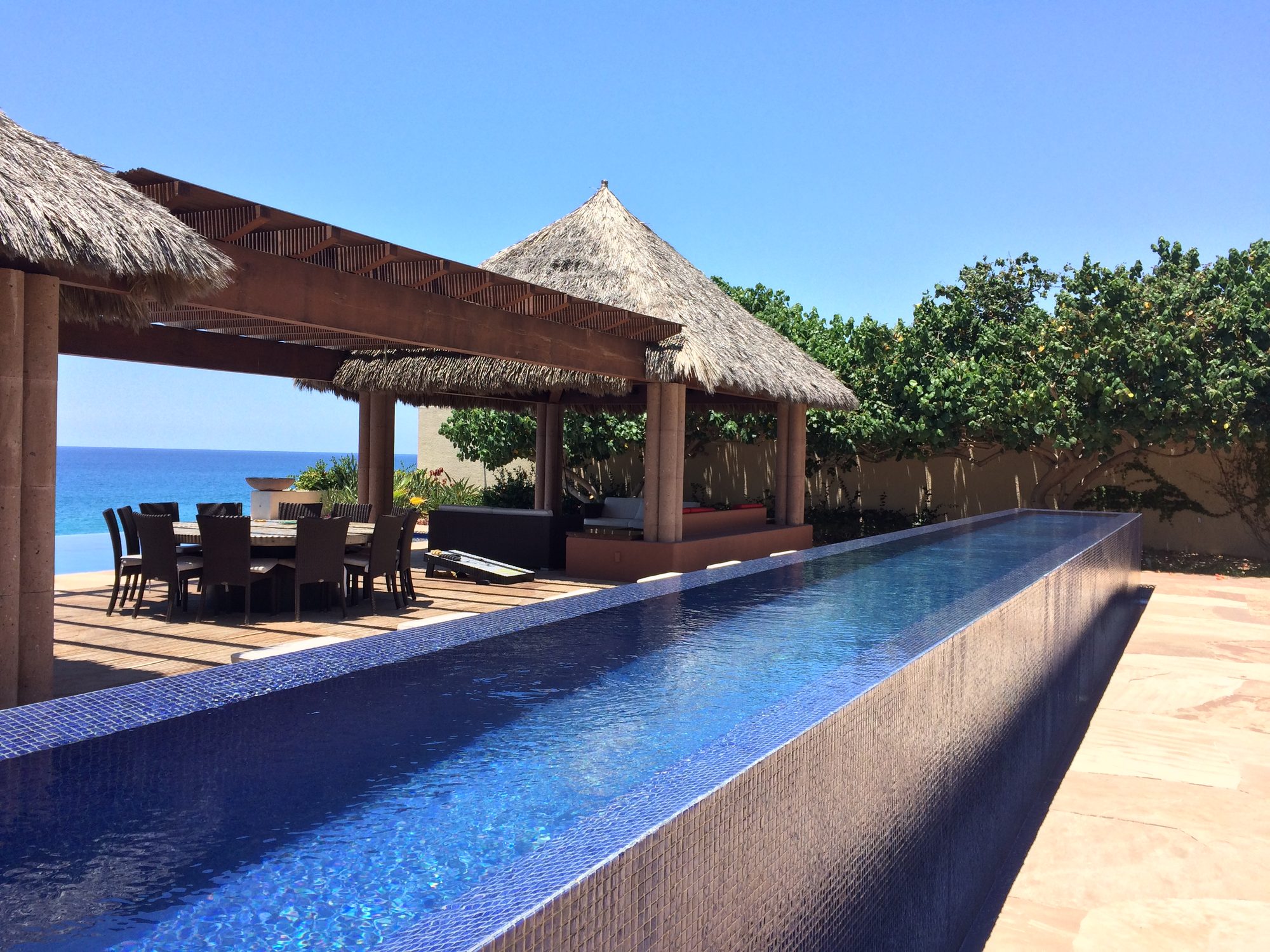 A villa infinity pool running toward the sea in Los Cabos, with a palapa-shaded dining table beside it