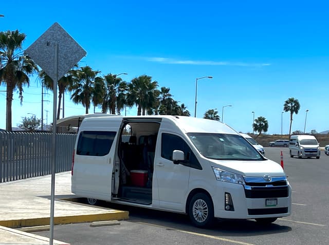 Van privada de Cabo Travel Boutique esperando con la puerta abierta, entre palmeras, en el aeropuerto de Los Cabos