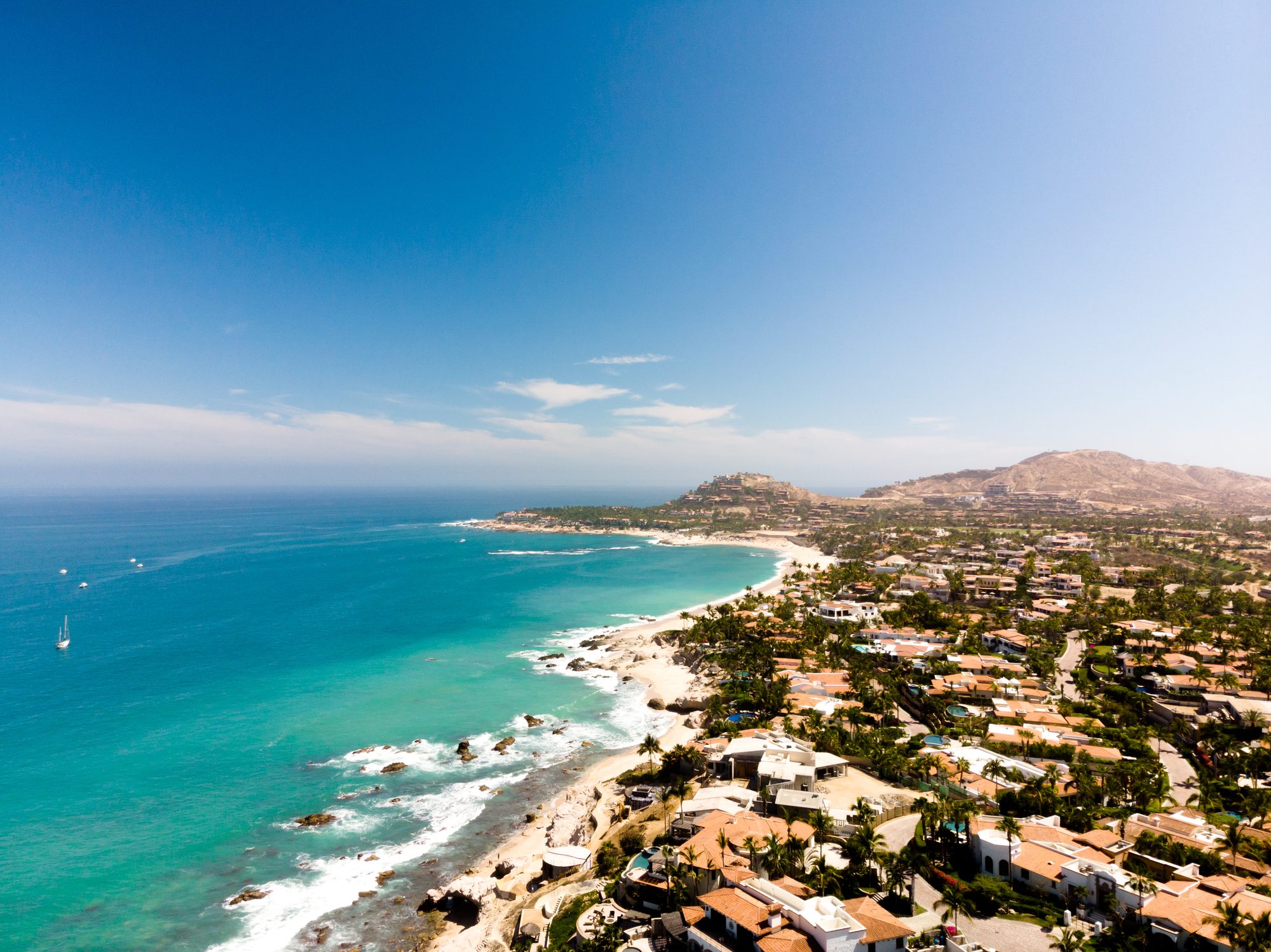 Aerial view of the Los Cabos coastline — turquoise water breaking on a rocky shore with hillside homes and desert mountains behind