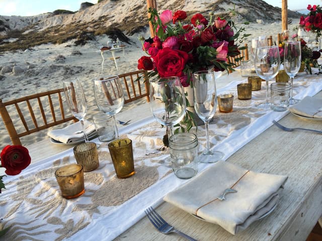 Mesa larga montada junto a la playa con flores, copas y velas al atardecer