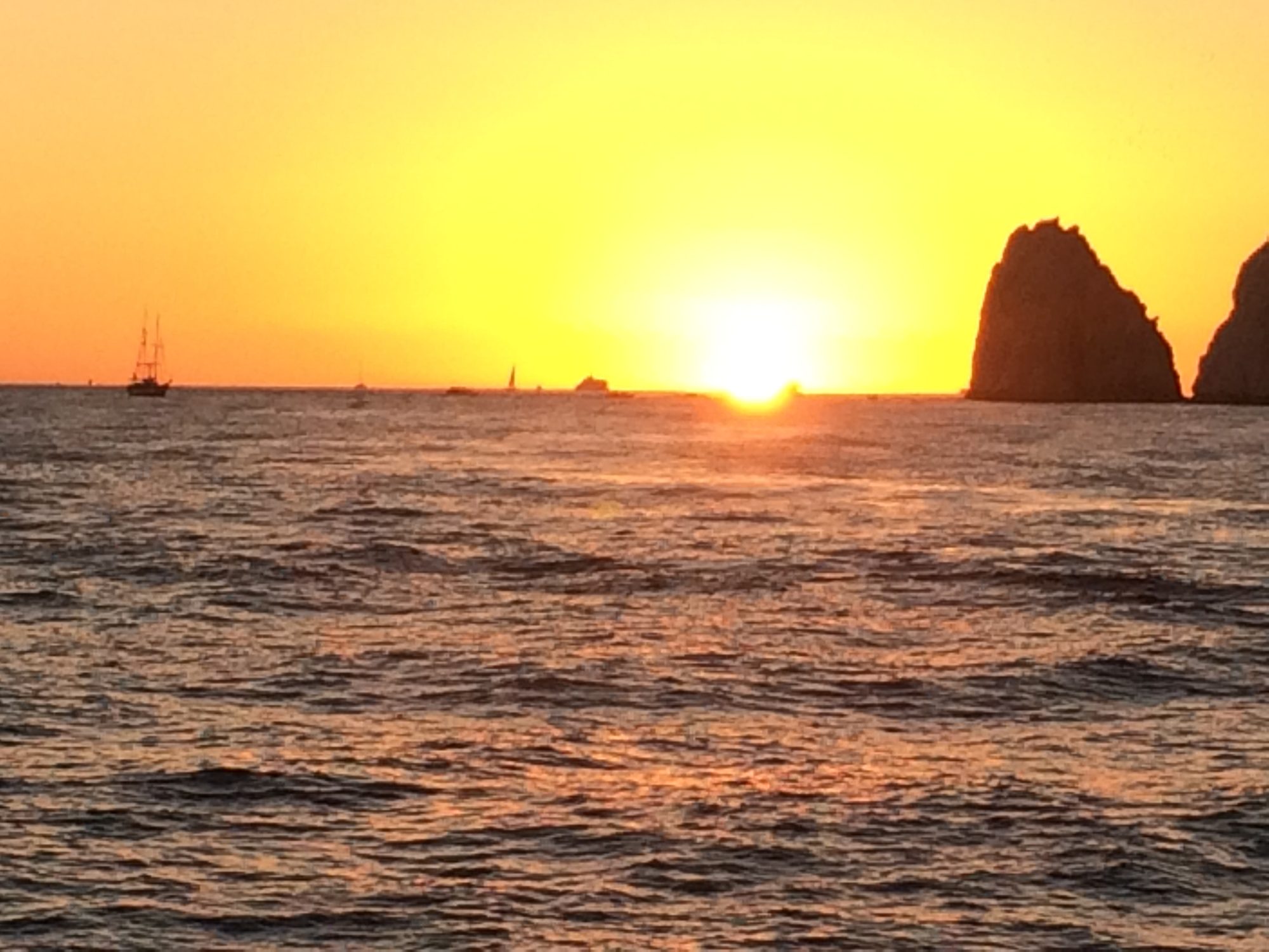 Sunset over the water off Los Cabos, with the Land's End rock formation on the horizon and boats in silhouette